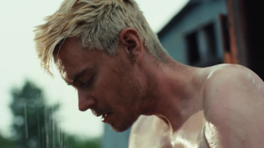 A shirtless man with bleached blonde hair washes himself under an outdoor shower in slow motion