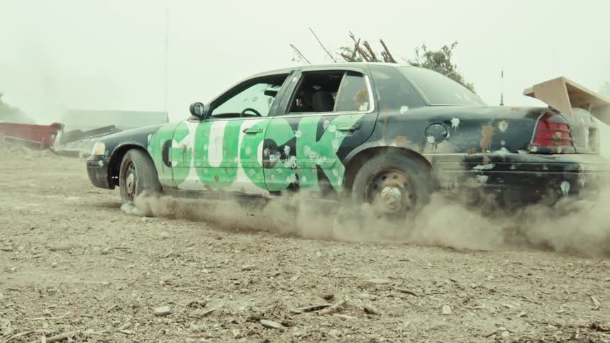 A beat-up car with graffiti spray painted on the side does a burnout in slow motion, kicking up a massive cloud of dirt in a junkyard