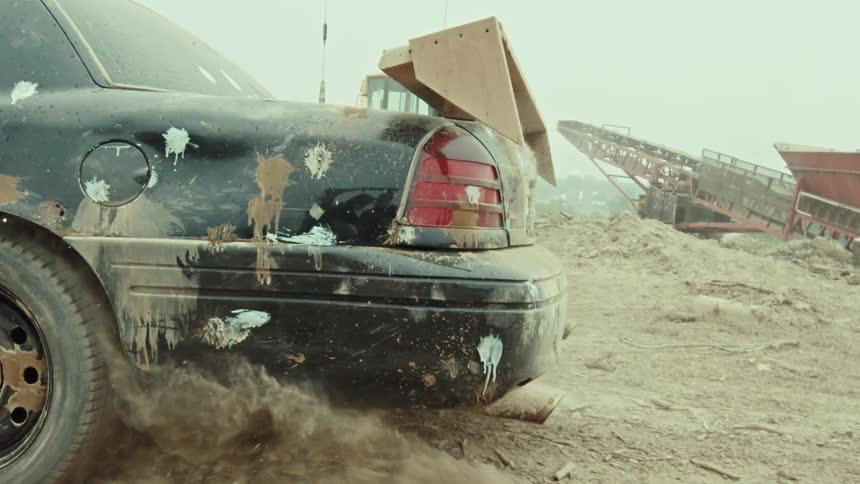 Slow motion shot of a muddy demolition derby car spinning its wheels, kicking up dirt and dust as its exhaust pipe breaks off