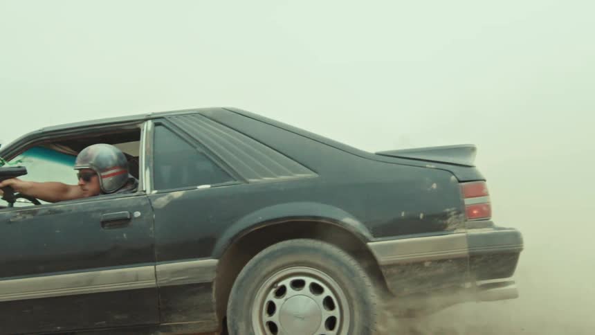 A man in a helmet and sunglasses aims a paintball gun from the window of a black car as it drives through a dusty wasteland in slow motion