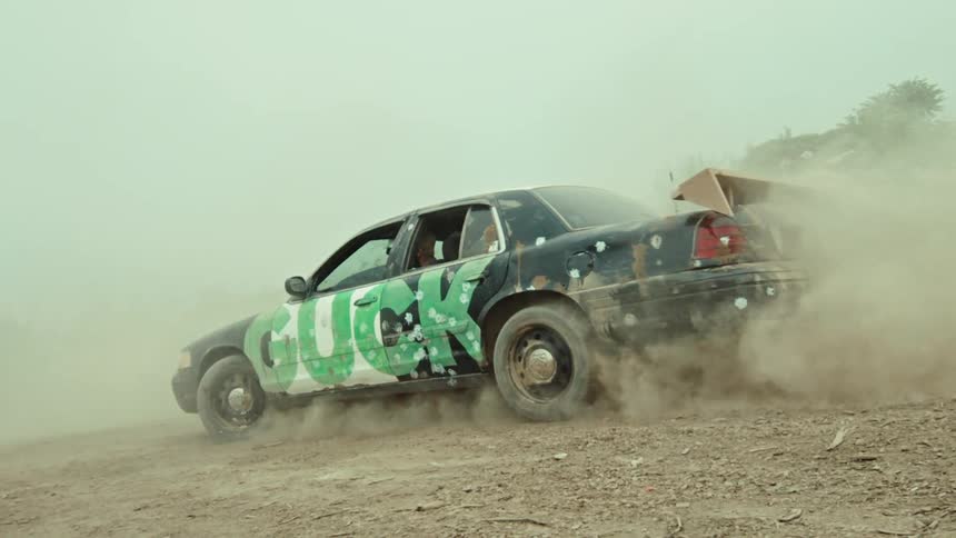 A beat-up, graffiti-covered car with bullet holes kicks up a cloud of dust as it spins its wheels and drives away in slow motion
