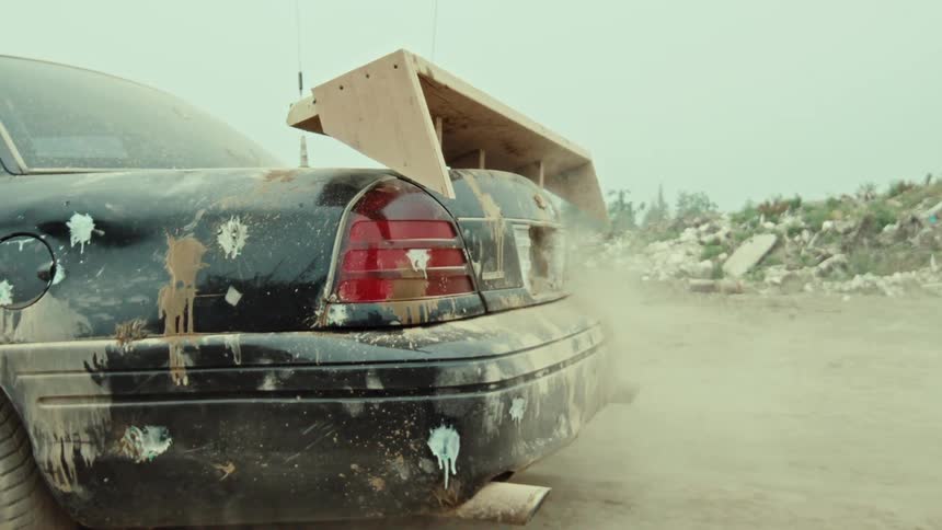 Smoke billows from the exhaust of a grimy, custom-modified black car idling in a desolate, post-apocalyptic junkyard