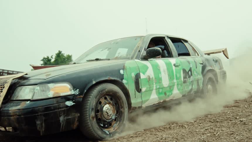 Slow motion footage shows a man in a custom derby car kicking up a cloud of dust as he drives through a rural junkyard