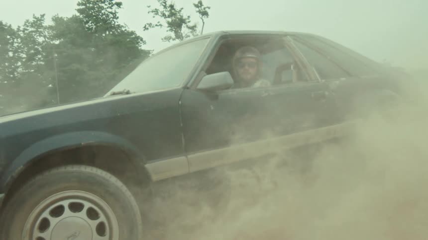 A driver in a helmet and sunglasses does a burnout in his black sports car, kicking up a massive cloud of dust in slow motion