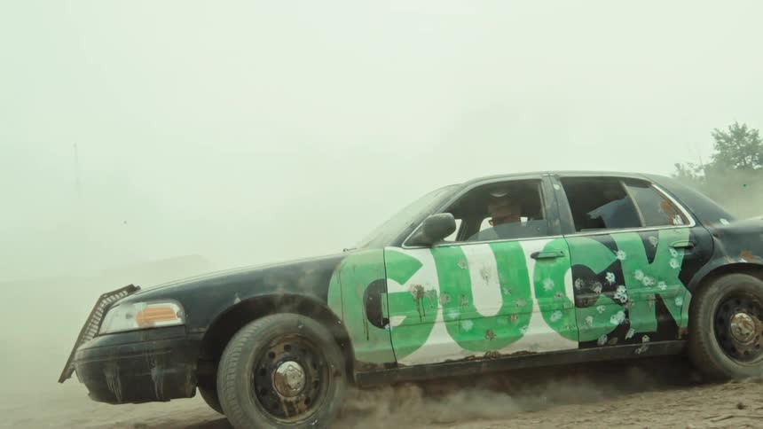 A man in a beat-up car with graffiti on the side does a burnout, kicking up dust and smoke in slow motion
