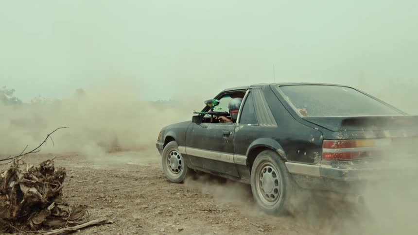 Dust swirls in slow motion around a black car as a man with a paintball gun takes aim in a desolate field