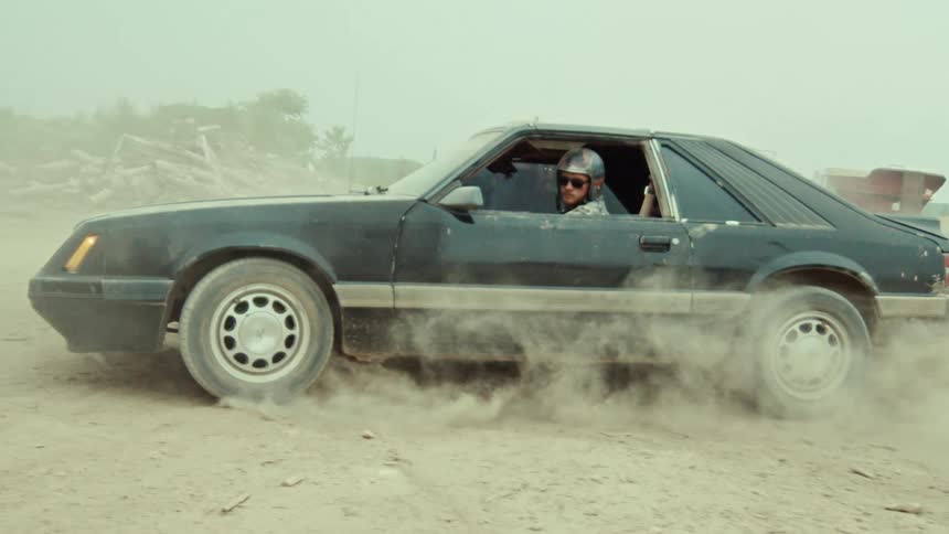 In slow motion, a man wearing a helmet performs a burnout in a vintage black muscle car, engulfing the vehicle in a thick cloud of dust