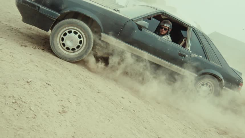 Slow motion side view of a man in a black Ford Mustang doing a burnout on a dirt road, kicking up a cloud of dust
