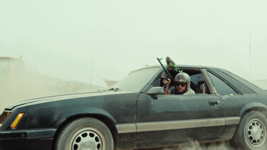 A man in a helmet aims a paintball gun from a black car doing a burnout, kicking up clouds of dust in slow motion