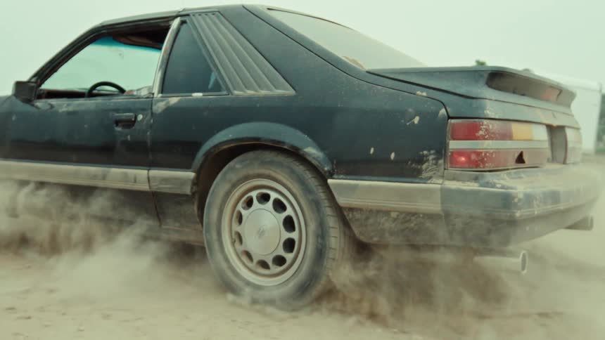 A close up slow motion shot of a vintage black Ford Mustang doing a burnout, its wheels spinning and kicking up dust on a dirt road