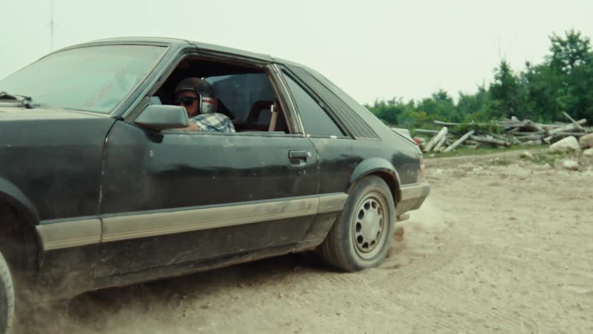 A man wearing a helmet performs a slow-motion burnout in an old black sports car, spinning the back tire and kicking up dust on a dirt road