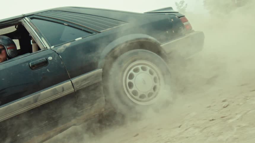A driver in a helmet does a burnout in a black muscle car on a dirt hill, kicking up a cloud of dust in slow motion