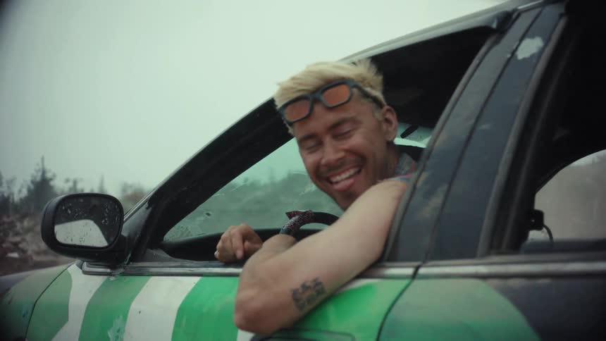 A smiling man in a demolition derby car looks at the camera before speeding off and kicking up a cloud of dust in slow motion