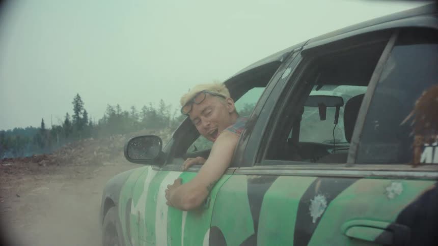 A man with bleached blonde hair hangs out of a moving car in slow motion as it kicks up dust on a dirt road