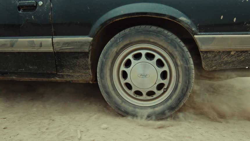 A close-up of a car's rear wheel spinning in slow motion on a dirt road, kicking up a cloud of dust