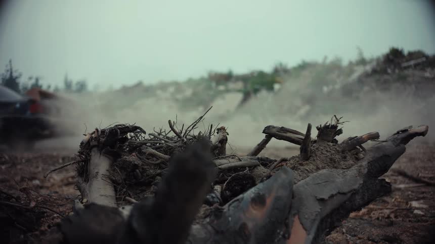 A car covered in green graffiti drives fast through a desolate, rubble-strewn landscape, kicking up a cloud of dust in slow motion