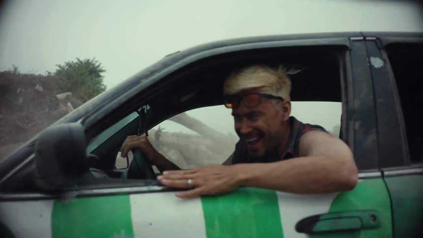 A wild-eyed man with bleached hair laughs maniacally as he peels out in his custom-painted car, kicking up a massive cloud of dust in a barren wasteland