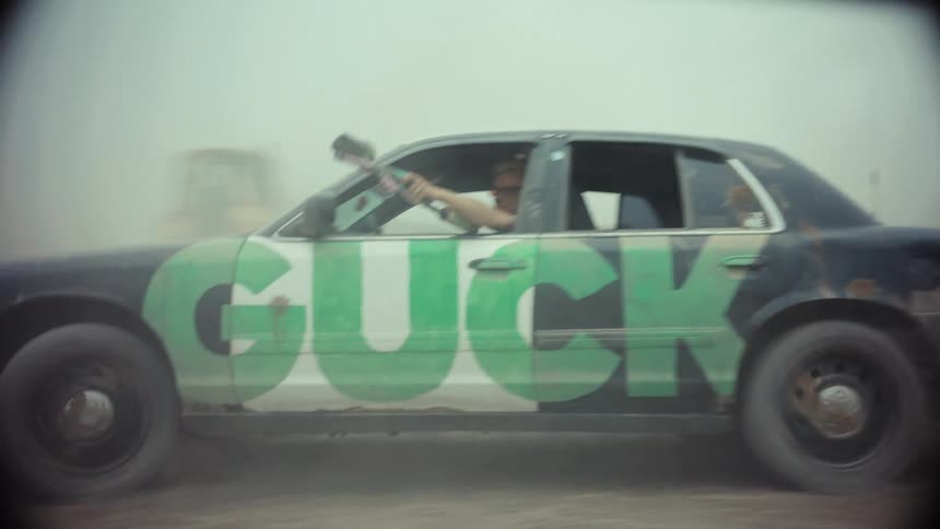 Slow motion tracking shot of a man leaning out of a custom car and shooting a paintball gun in a dusty, barren landscape