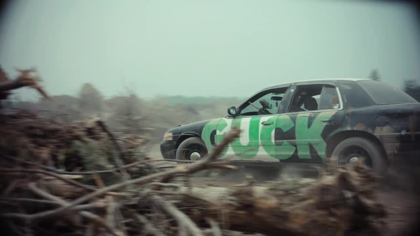 A black sedan with green graffiti speeds down a dirt road, kicking up dust in a desolate, debris-filled wasteland