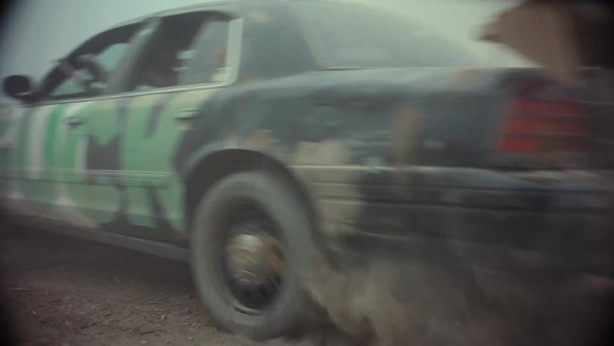 A gritty, close-up tracking shot shows a modified car with a paintball gunner speeding down a dusty dirt road, kicking up a massive cloud of dust