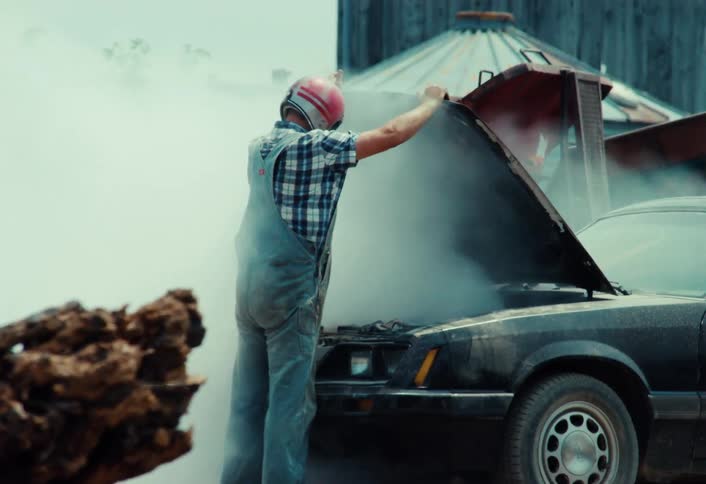 A frustrated man in a helmet and overalls kicks his smoking car in anger during a demolition derby