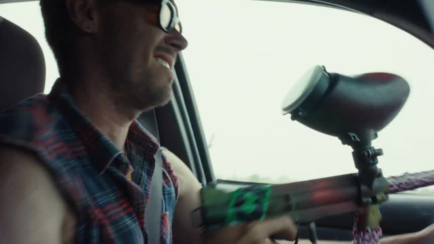 A man in sunglasses drives his car while shooting a paintball gun out of the passenger side window