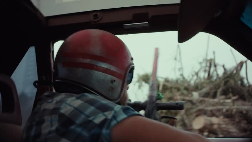 A man in a vintage red helmet and plaid shirt drives a car while holding a paintball gun in slow motion