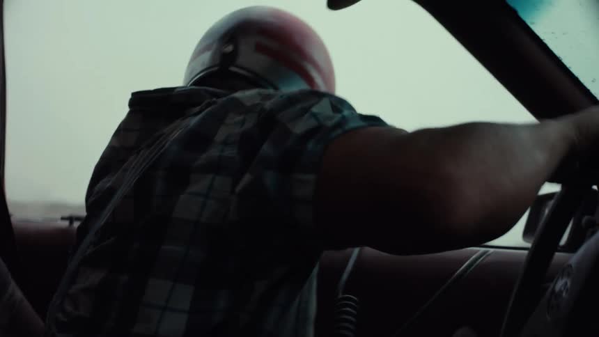 A slow-motion over-the-shoulder shot shows a man in a helmet shooting a paintball gun from a speeding car