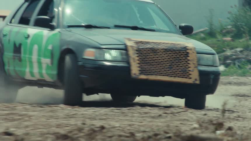 A modified race car with green graffiti drifts in slow motion along a dirt track, kicking up a large cloud of dust