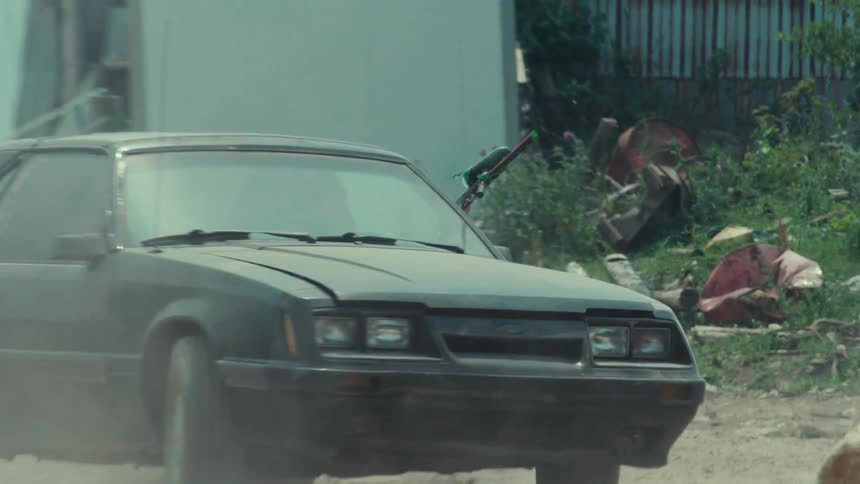 Two cars drive through a dusty junkyard as a passenger shoots a paintball gun from one of the vehicles in slow motion