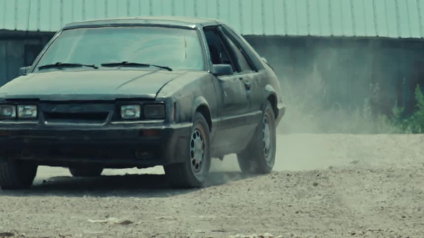 A weathered black vintage hatchback idles on a dirt road as dust kicks up from the rear tire in slow motion