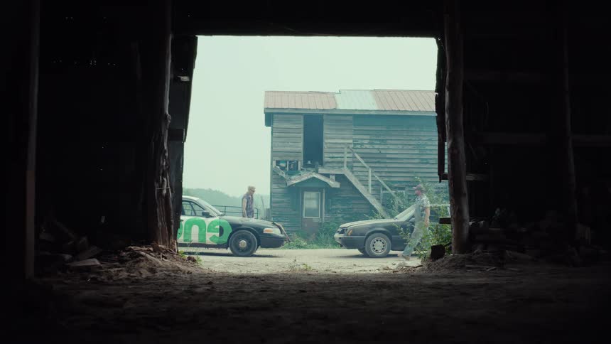 Viewed from a dark barn, two men meet between their cars in a rural yard before a heated argument escalates into a physical fight