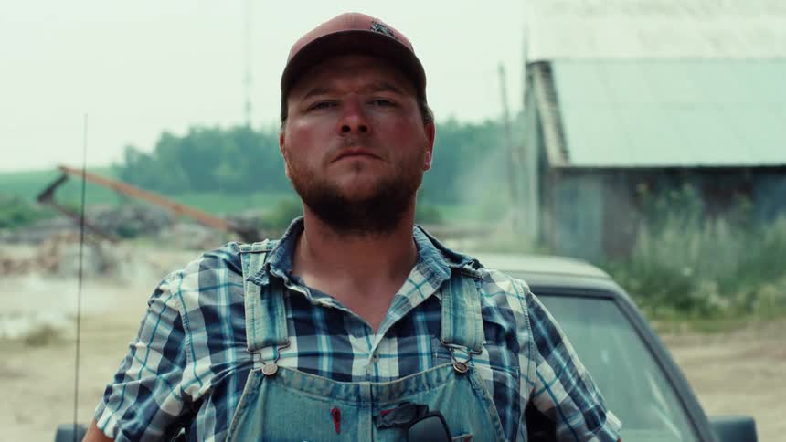 A tough-looking farmer in a baseball cap and overalls walks towards the camera, takes off his sunglasses, and stares with a serious expression in a rural setting