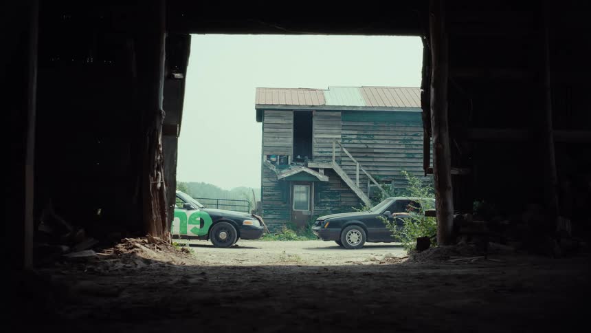 A tense face-off between two cars outside a rustic building, framed by the dark entrance of an old barn