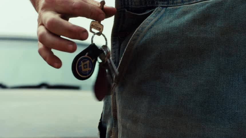 A man's weathered hand reaches into his denim overall pocket and pulls out a keychain in slow motion