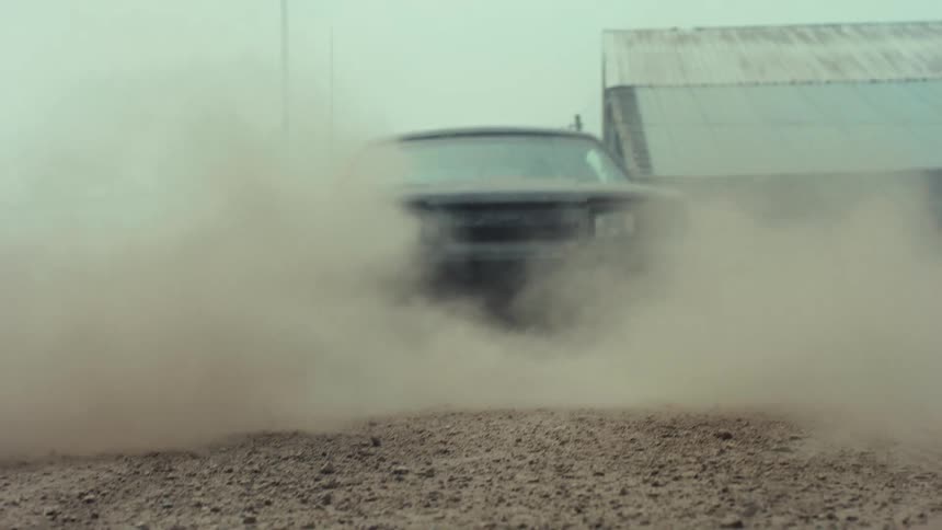 A low-angle view of a vintage car doing a reverse burnout, creating a massive dust cloud on a dirt road