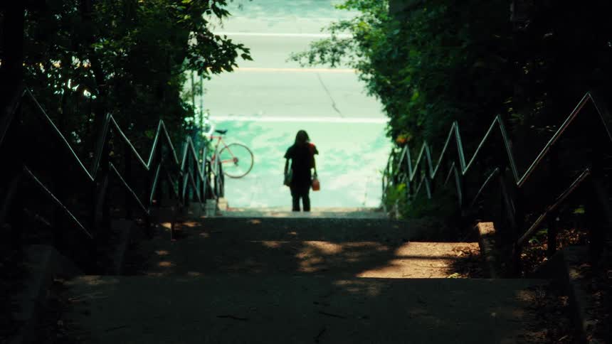 A silhouetted person on a skateboard practices tricks at the bottom of a shaded staircase before walking away into the summer sunlight