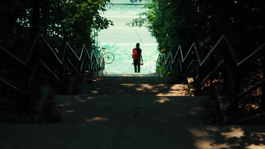 A view from the top of a dark, wooded staircase shows a person with a red backpack standing in a bright green bike lane as traffic passes by