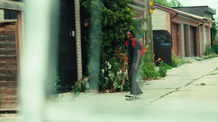 A young man with a red backpack casually skateboards through a gritty urban alleyway in slow motion