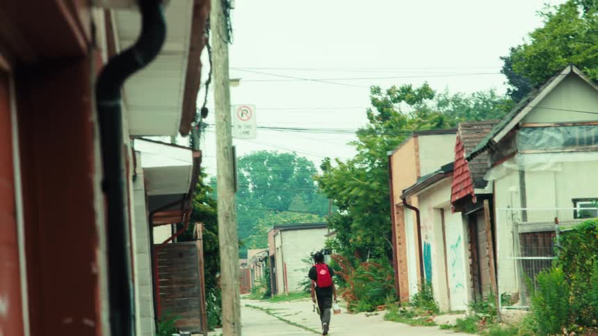 A person with a bright red backpack walks down a rundown urban alleyway carrying a skateboard