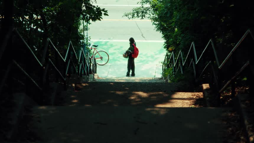 From the top of a shady staircase, a person with a backpack and skateboard waits by a bicycle next to a green bike lane