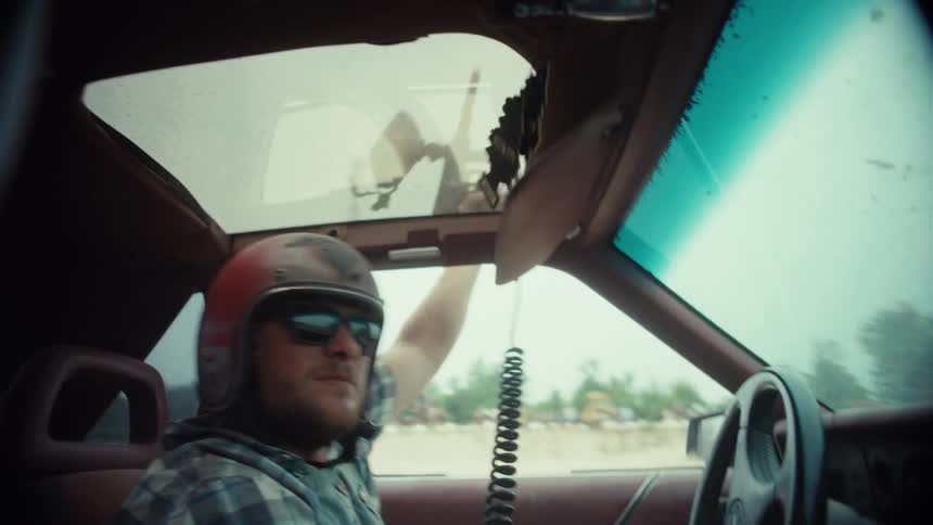 A man in a helmet drives a demolition derby car while shooting a paintball gun out the window