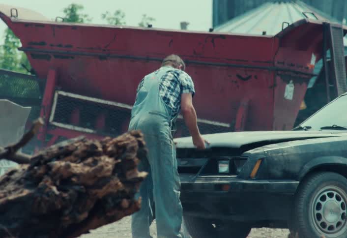 A frustrated man in overalls gets out of his broken-down muscle car, inspects the engine, and gives up in exasperation at a junkyard