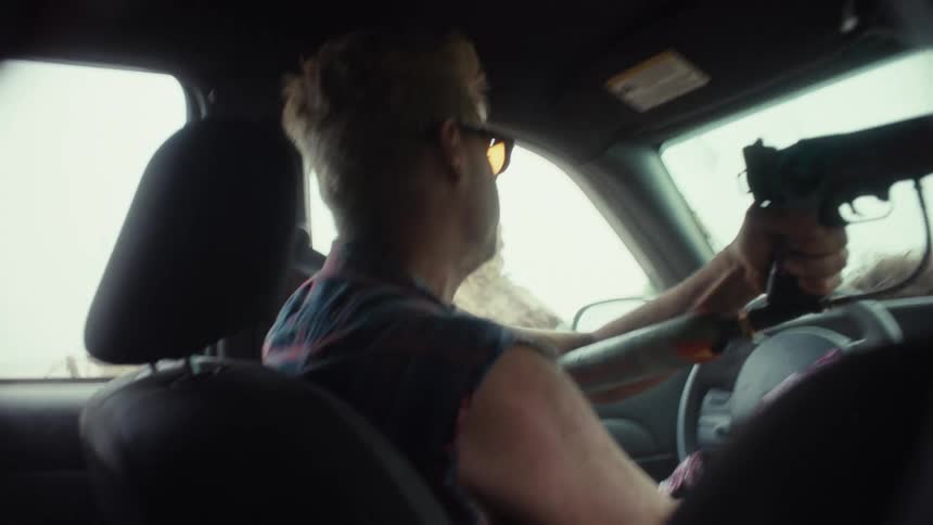 A rebellious man with bleached blonde hair drives through a hazy landscape, aiming a paintball gun out of his car window