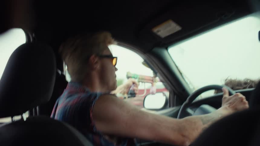 Energetic man in a sleeveless plaid shirt and sunglasses drives and shoots a paintball gun from his car window in slow motion