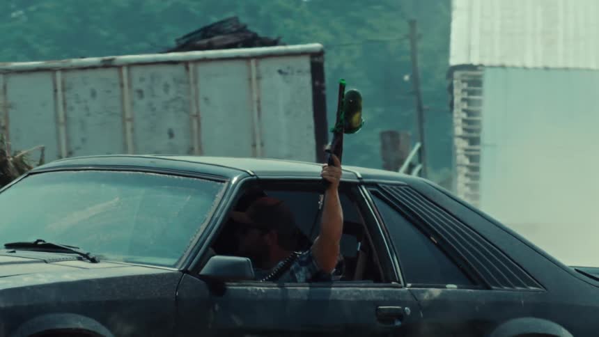 Driver of a classic black sports car holds a paintball gun aloft as smoke from a burnout fills the air in slow motion