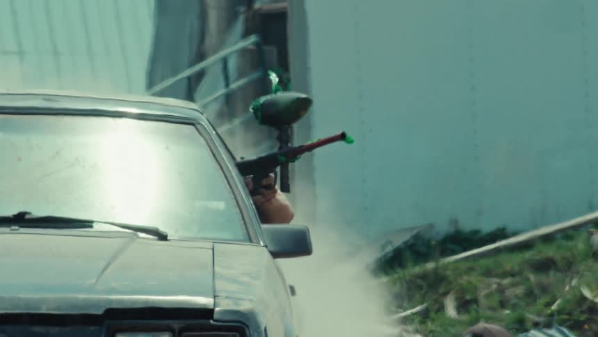 Slow motion close-up of a paintball player taking cover behind a car and aiming their marker at an opponent during an intense game