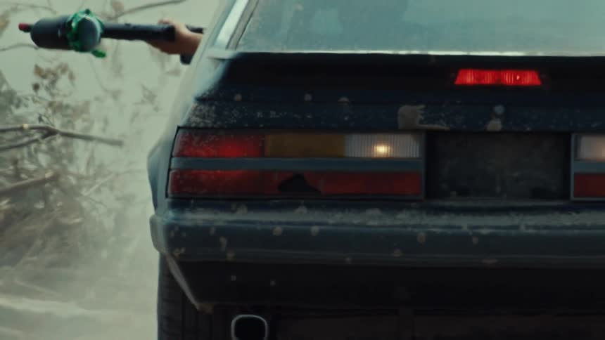 A slow zoom out from the broken taillight of a dirty vintage car reveals a passenger aiming a rocket launcher out the window
