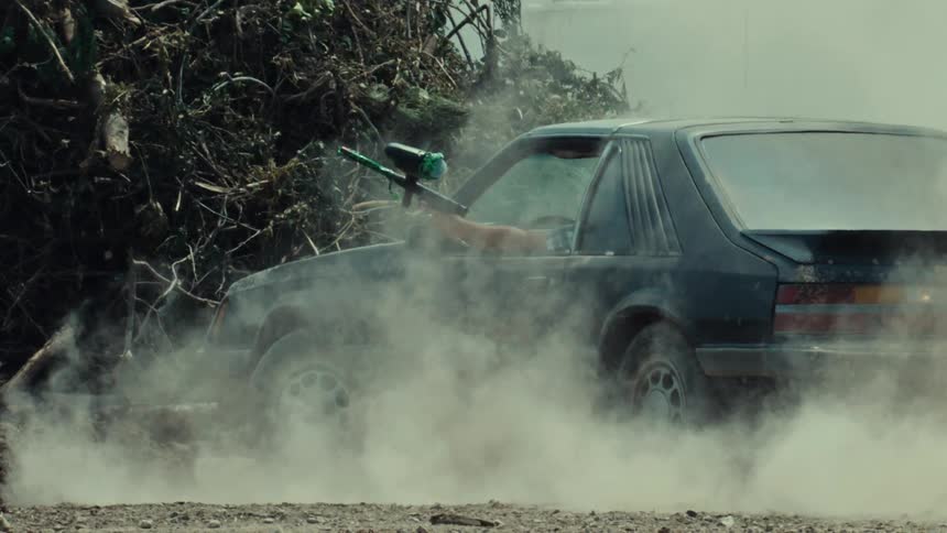 A man shoots a paintball gun in slow motion from the window of a car surrounded by smoke and dust