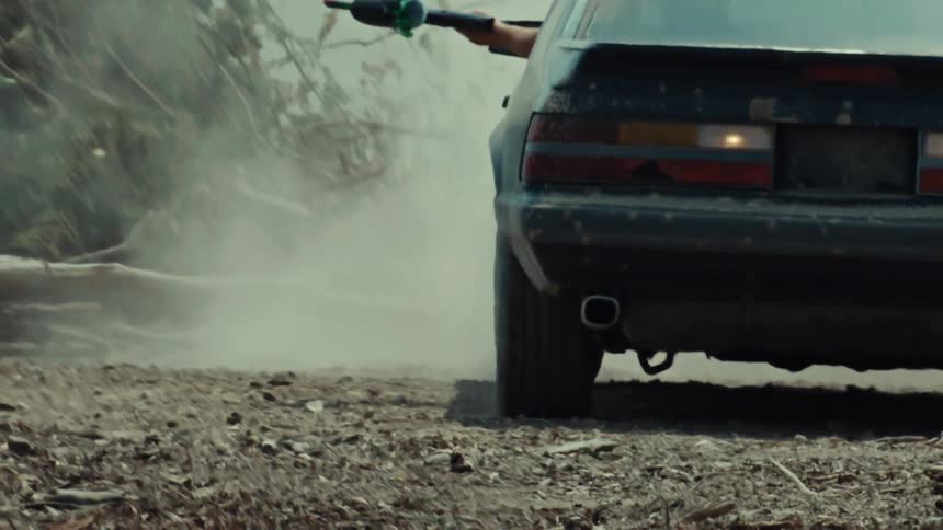 A person aims a weapon out of a beat-up black car window as dust billows around in slow motion